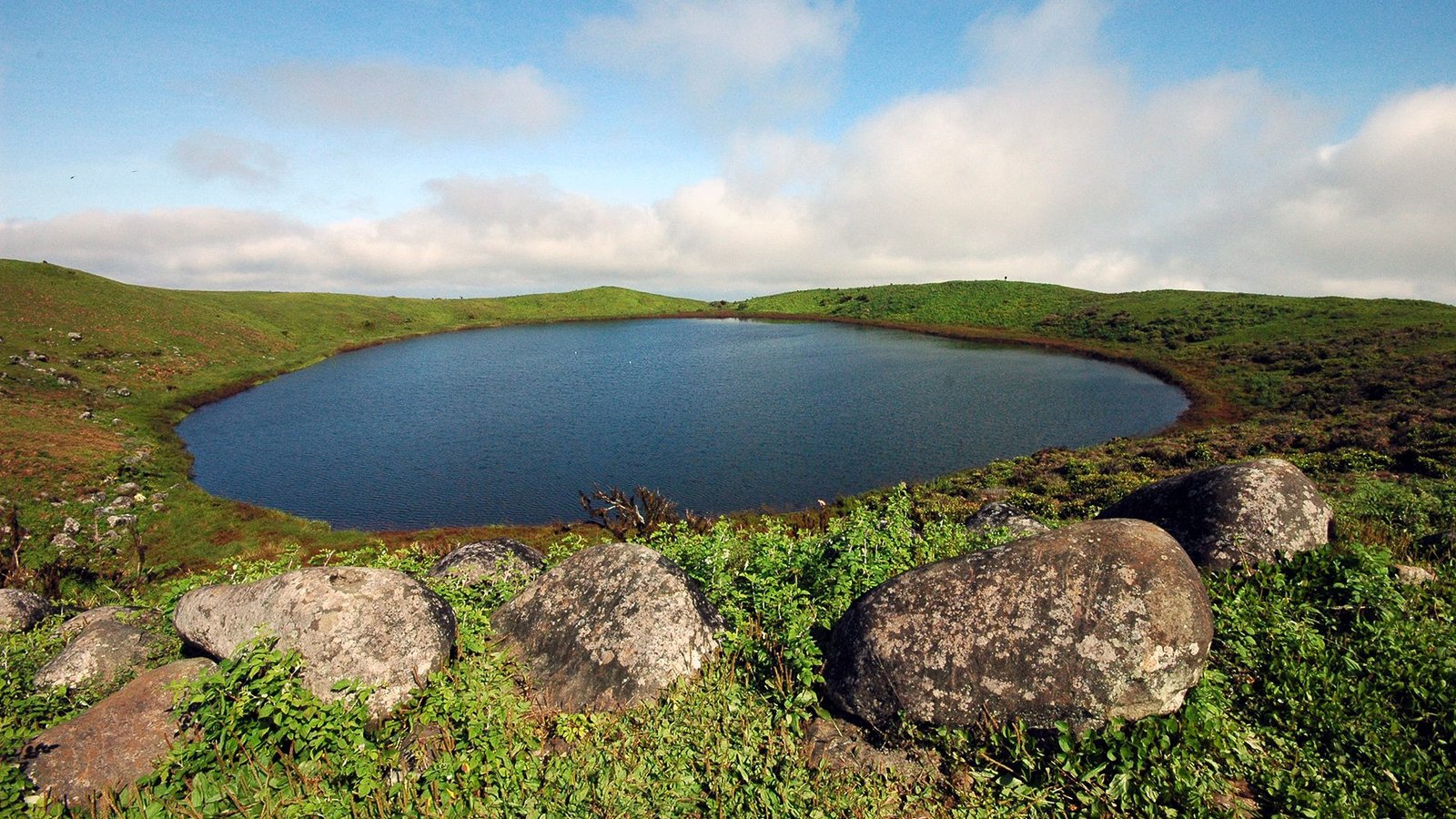 Laguna circular en un cráter volcánico de las islas Galápagos, rodeada de colinas verdes y grandes rocas en primer plano.