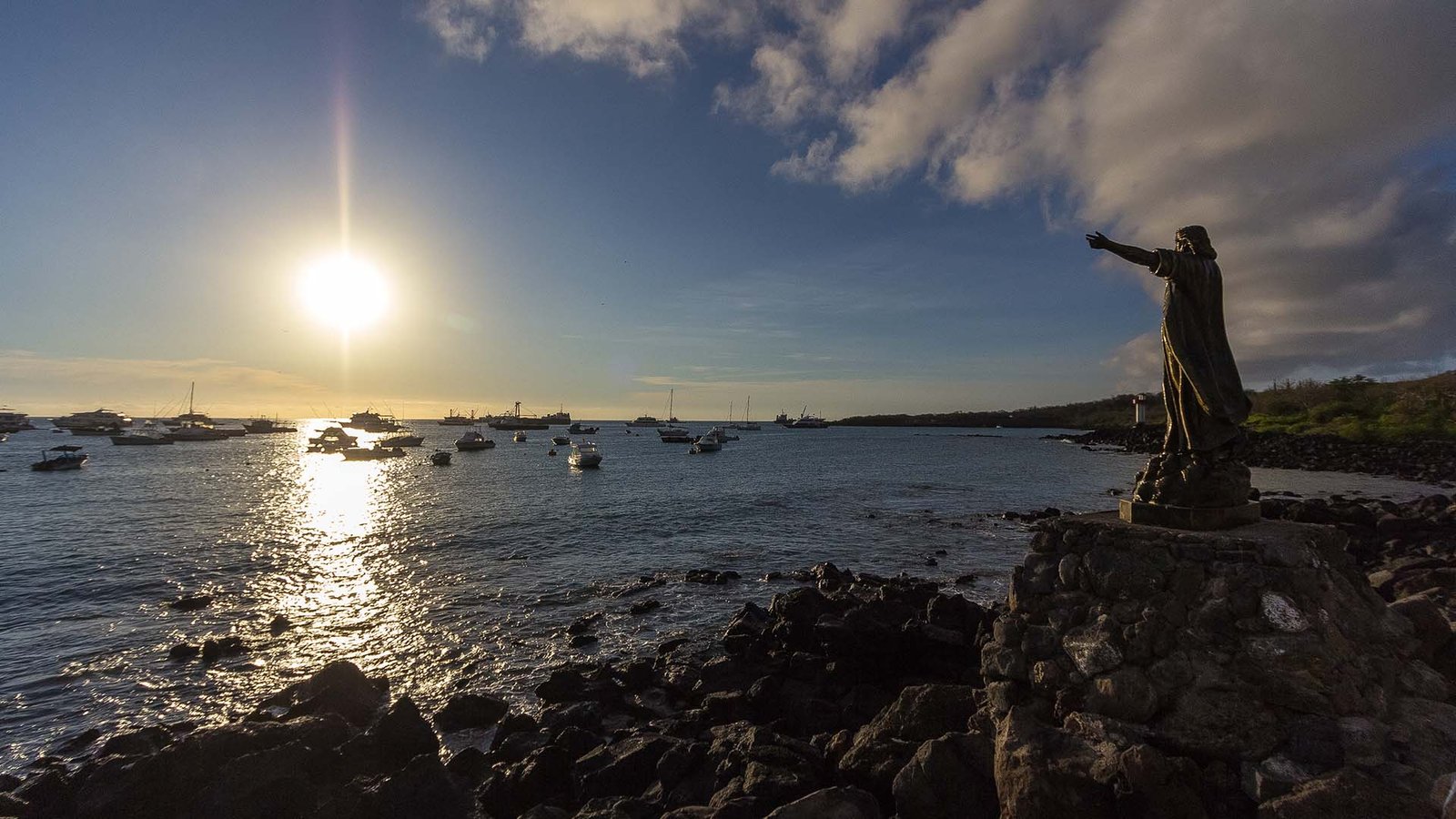 Statue of Charles Darwin overlooking the ocean at sunset in Puerto Ayora, Galápagos, with boats anchored in the bay.