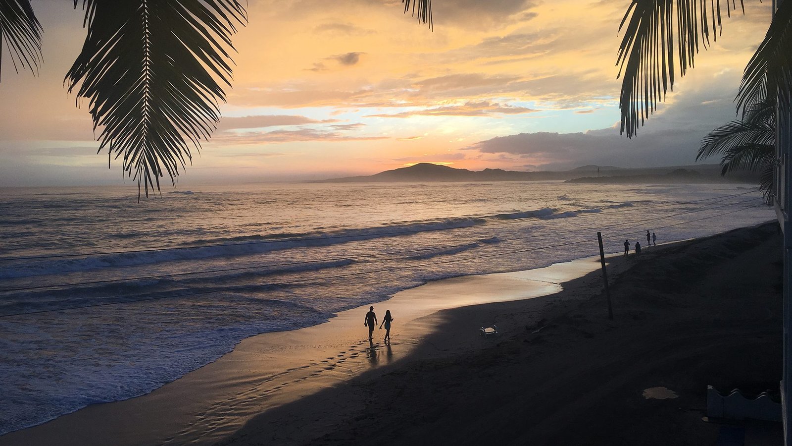 Couple walking along the Pacific Coast of Ecuador at sunset, framed by palm trees with golden skies and gentle waves.